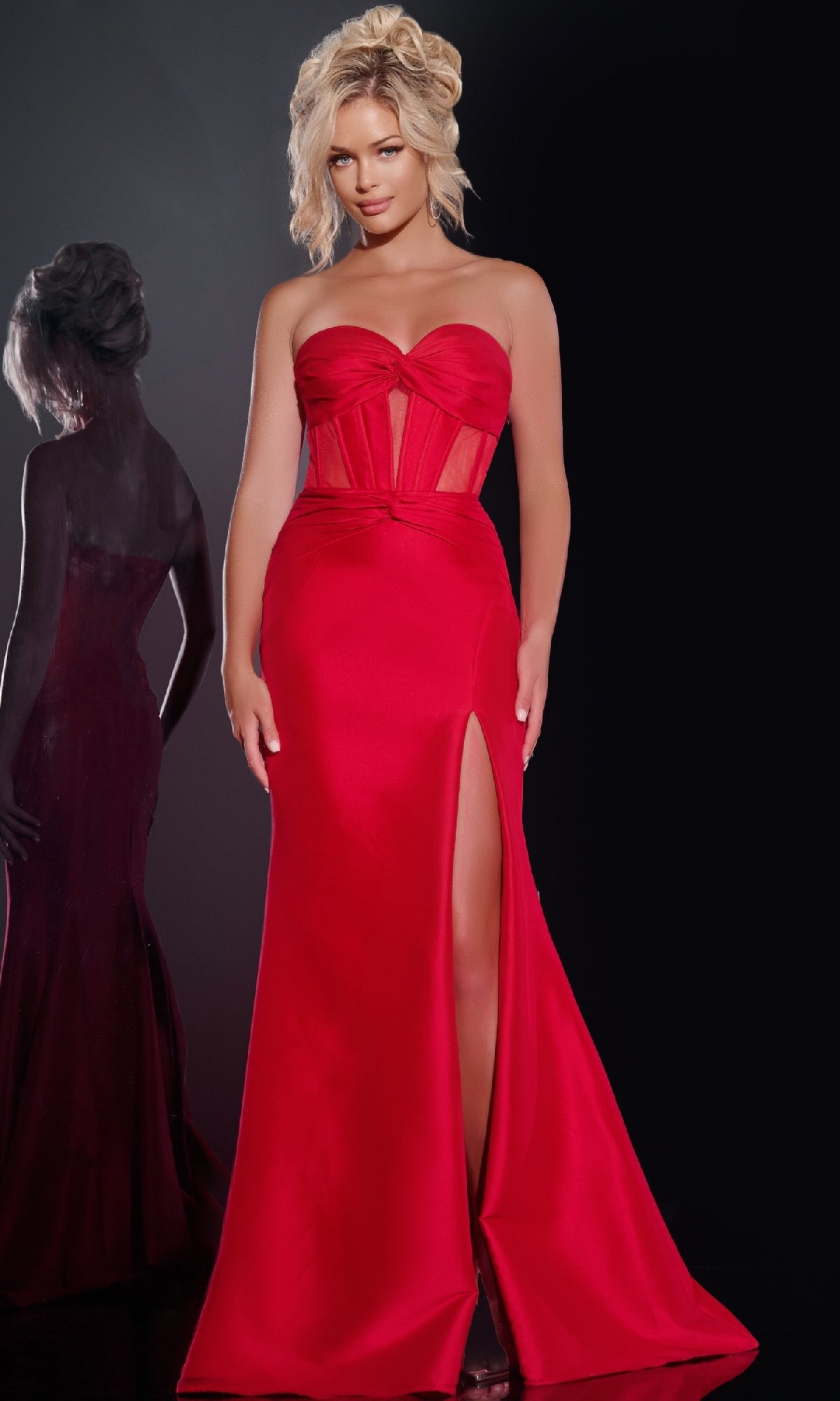 A woman poses against a dark background, her reflection visible on the left, wearing the Jovani 42375 Long Prom Dress: a strapless red gown with a corset bodice, floor-length skirt, and high slit.